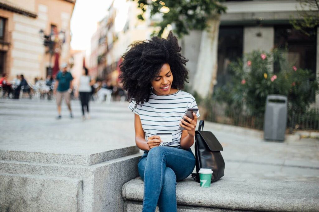 Smiling woman sitting outdoors holding a phone and credit card, with a backpack and coffee cup beside her.