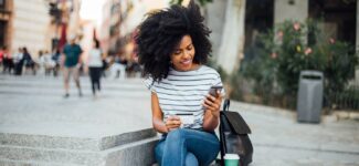 Smiling woman sitting outdoors holding a phone and credit card, with a backpack and coffee cup beside her.