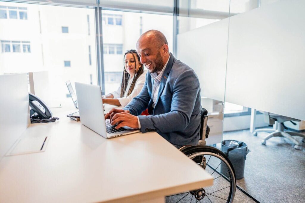 Man in a wheelchair working on a laptop at an office desk, smiling, with a female colleague beside him.