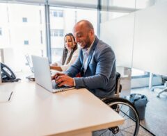 Man in a wheelchair working on a laptop at an office desk, smiling, with a female colleague beside him.