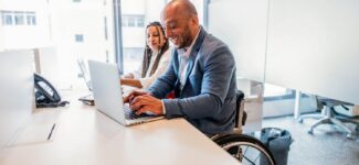 Man in a wheelchair working on a laptop at an office desk, smiling, with a female colleague beside him.