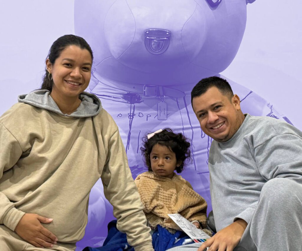 Smiling family with a young child sits together in front of a large teddy bear backdrop.