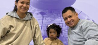 Smiling family with a young child sits together in front of a large teddy bear backdrop.