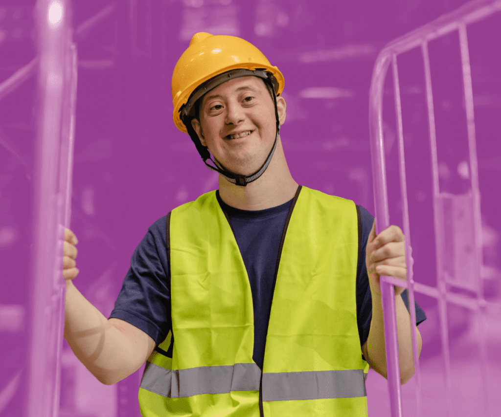 Smiling worker wearing a yellow safety vest and hard hat, standing between metal barriers in an industrial setting.