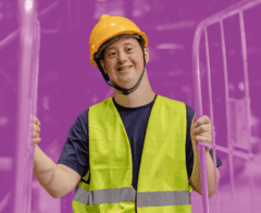 Smiling worker wearing a yellow safety vest and hard hat, standing between metal barriers in an industrial setting.