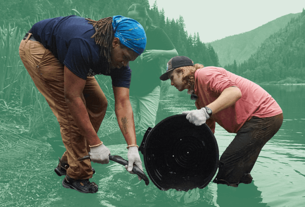 Two people working together in a river, one holding a net and the other pouring water from a large container, with forested mountains in the background.