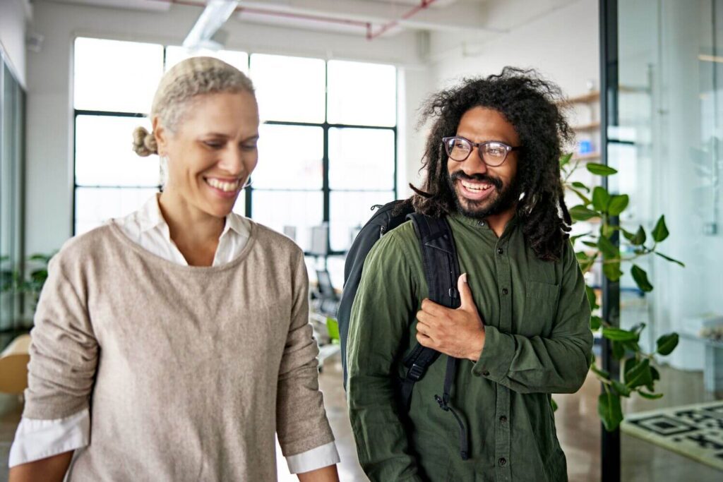 Two colleagues smiling and walking through a modern office space, engaged in a friendly conversation.