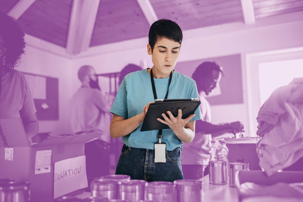 Volunteer using a tablet to manage inventory at a donation center, with canned goods and other volunteers working in the background.