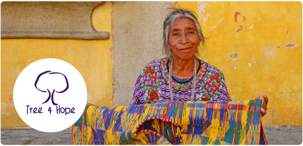 Smiling woman in traditional clothing holding colorful woven fabric in front of a yellow wall, with the Tree 4 Hope logo on the left.
