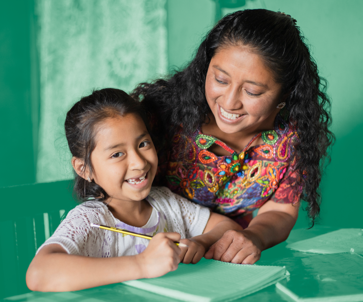 Smiling woman helping a young girl write in a notebook at a table, both wearing colorful clothing.