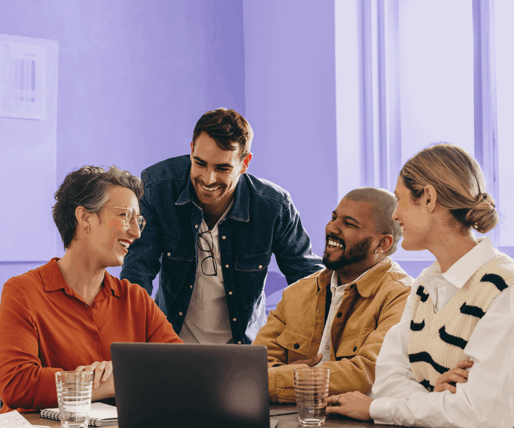 Group of four coworkers smiling and chatting around a laptop in a brightly lit office.