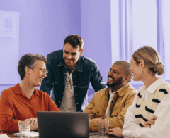 Group of four coworkers smiling and chatting around a laptop in a brightly lit office.