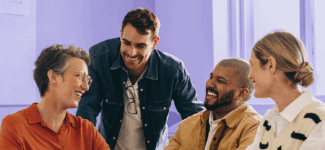 Group of four coworkers smiling and chatting around a laptop in a brightly lit office.