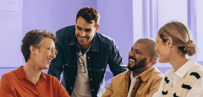 Group of four coworkers smiling and chatting around a laptop in a brightly lit office.