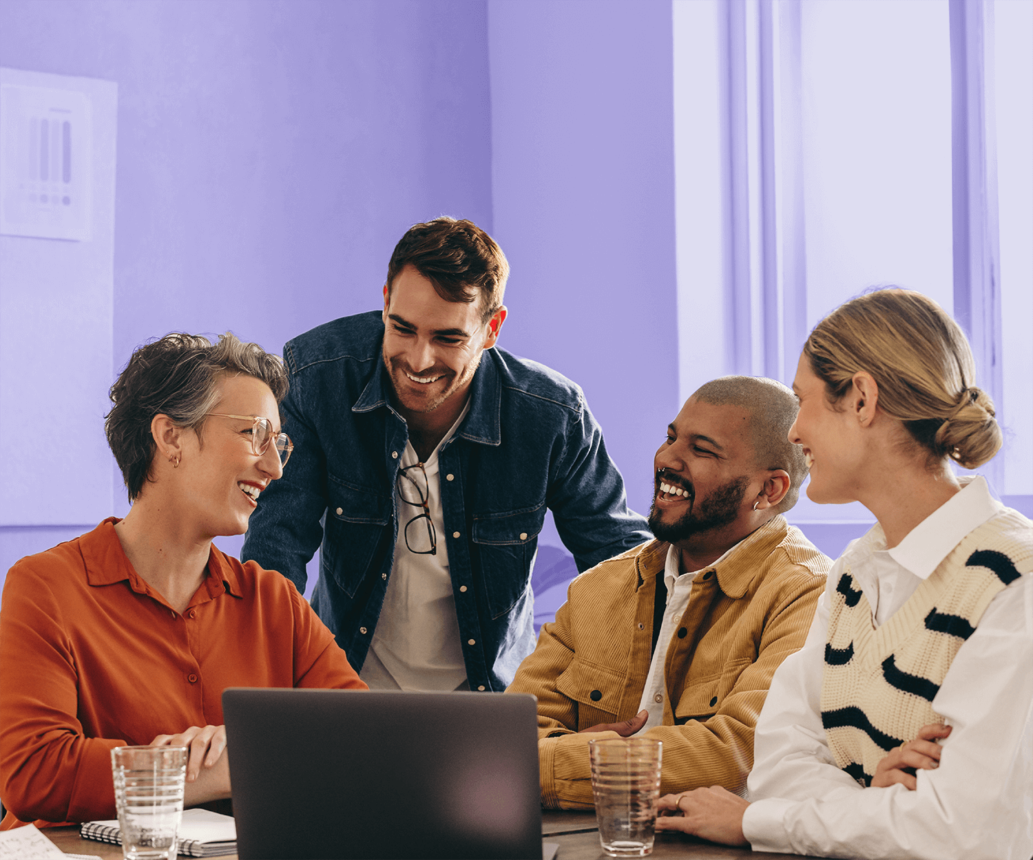 Group of four coworkers smiling and chatting around a laptop in a brightly lit office.