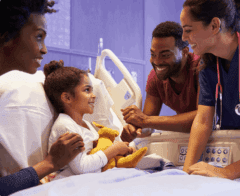 Smiling child in a hospital bed surrounded by supportive family members and a nurse.