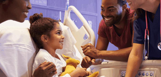 Smiling child in a hospital bed surrounded by supportive family members and a nurse.