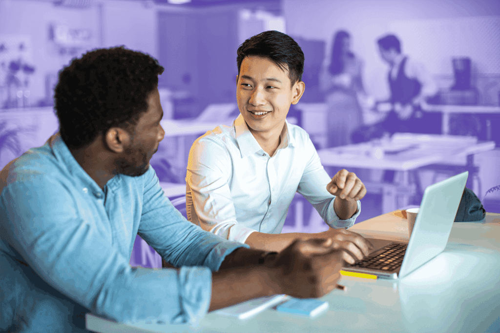 Two colleagues smiling and talking while working on a laptop in a modern office space.