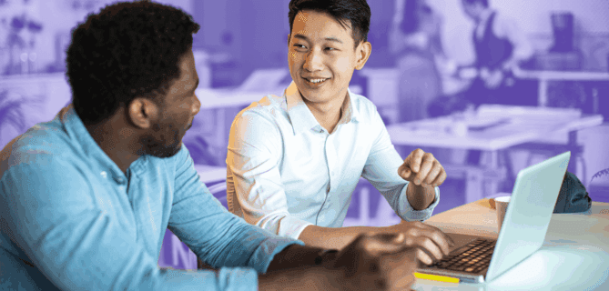Two colleagues smiling and talking while working on a laptop in a modern office space.
