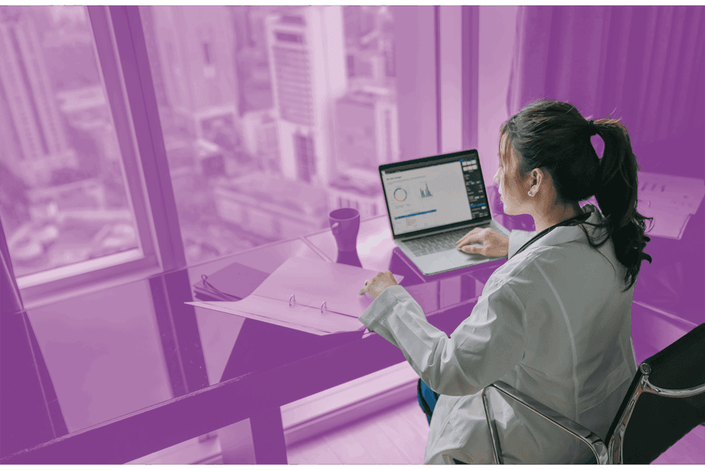 Woman in a lab coat working on a laptop with data charts in a high-rise office overlooking a cityscape.