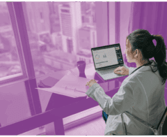 Woman in a lab coat working on a laptop with data charts in a high-rise office overlooking a cityscape.