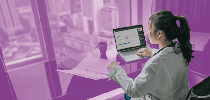 Woman in a lab coat working on a laptop with data charts in a high-rise office overlooking a cityscape.