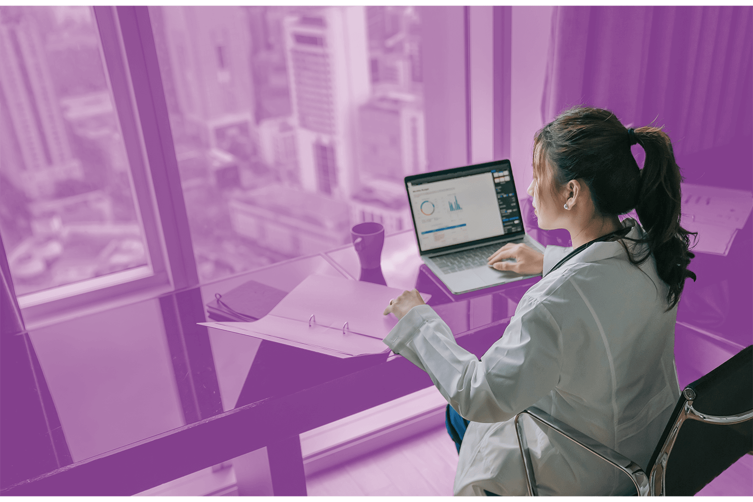 Woman in a lab coat working on a laptop with data charts in a high-rise office overlooking a cityscape.