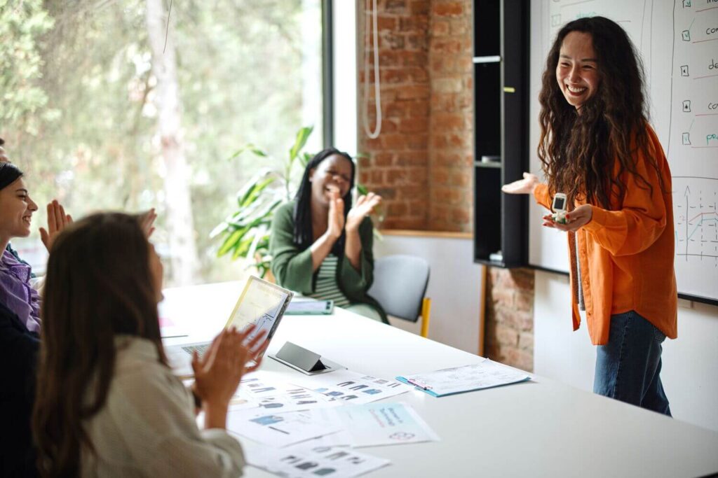 Woman presenting at a whiteboard while colleagues seated at a table applaud in a bright, modern office.