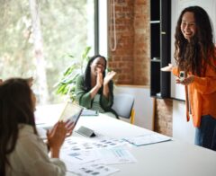 Woman presenting at a whiteboard while colleagues seated at a table applaud in a bright, modern office.