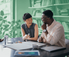 Two coworkers smiling and discussing documents at a table in a modern office with bookshelves.