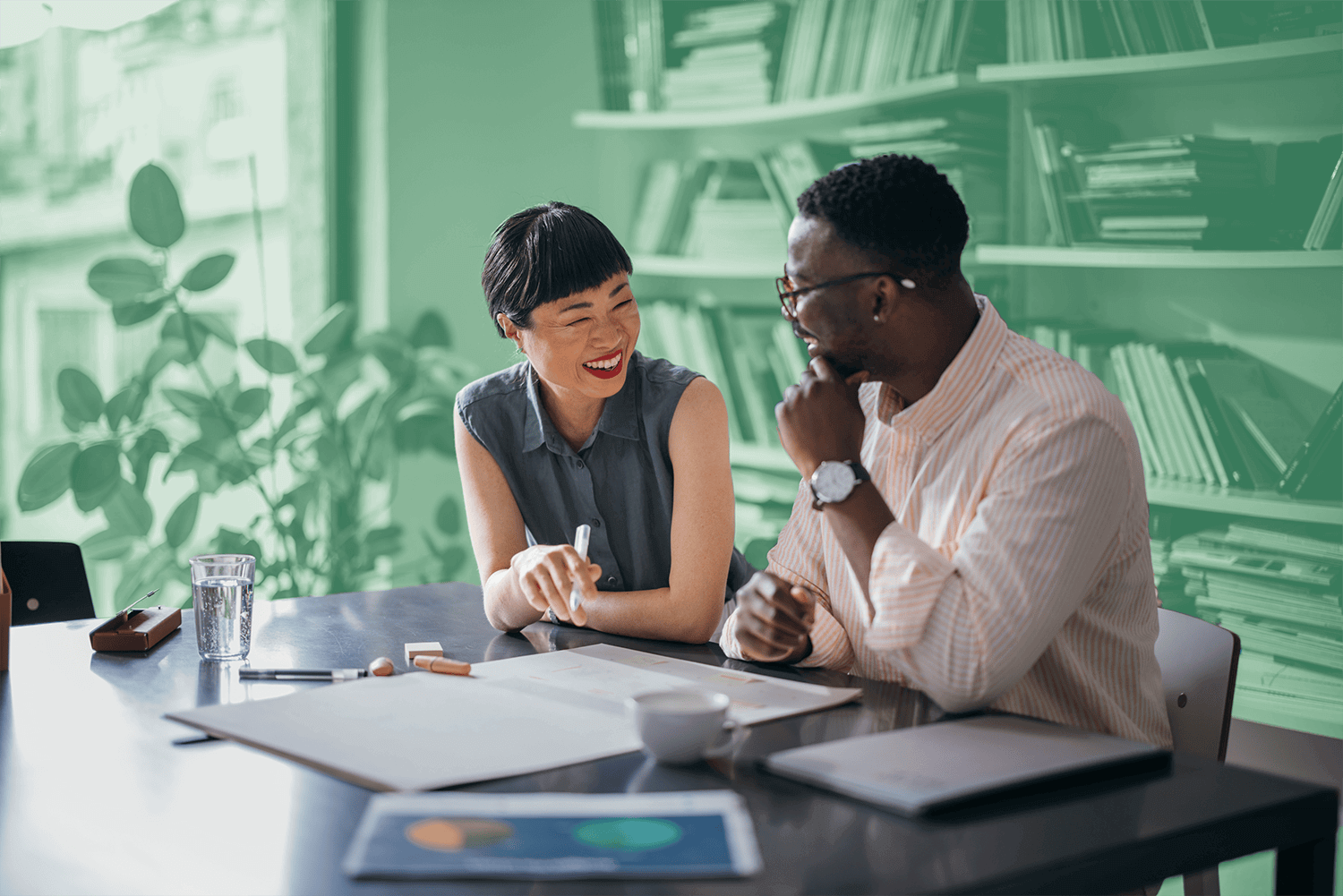 Two coworkers smiling and discussing documents at a table in a modern office with bookshelves.