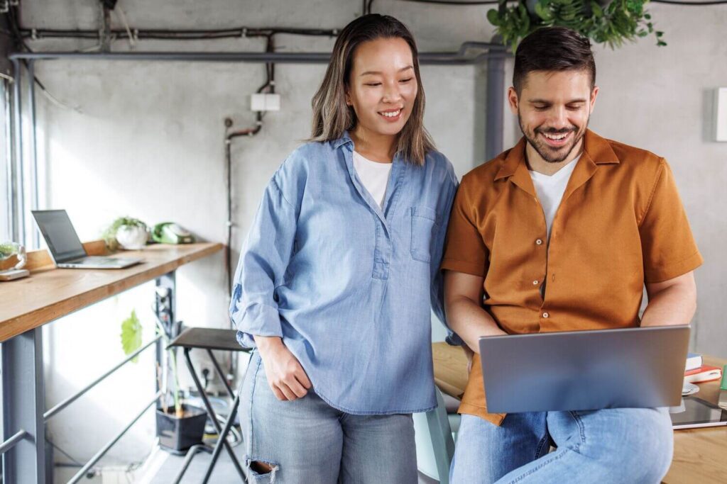Two colleagues smiling while looking at a laptop in a modern workspace.