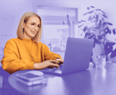 Smiling woman in a yellow sweater working on a laptop at a table in a bright home environment.