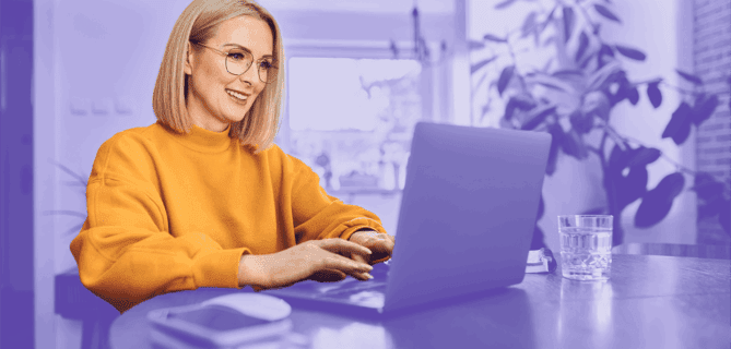 Smiling woman in a yellow sweater working on a laptop at a table in a bright home environment.