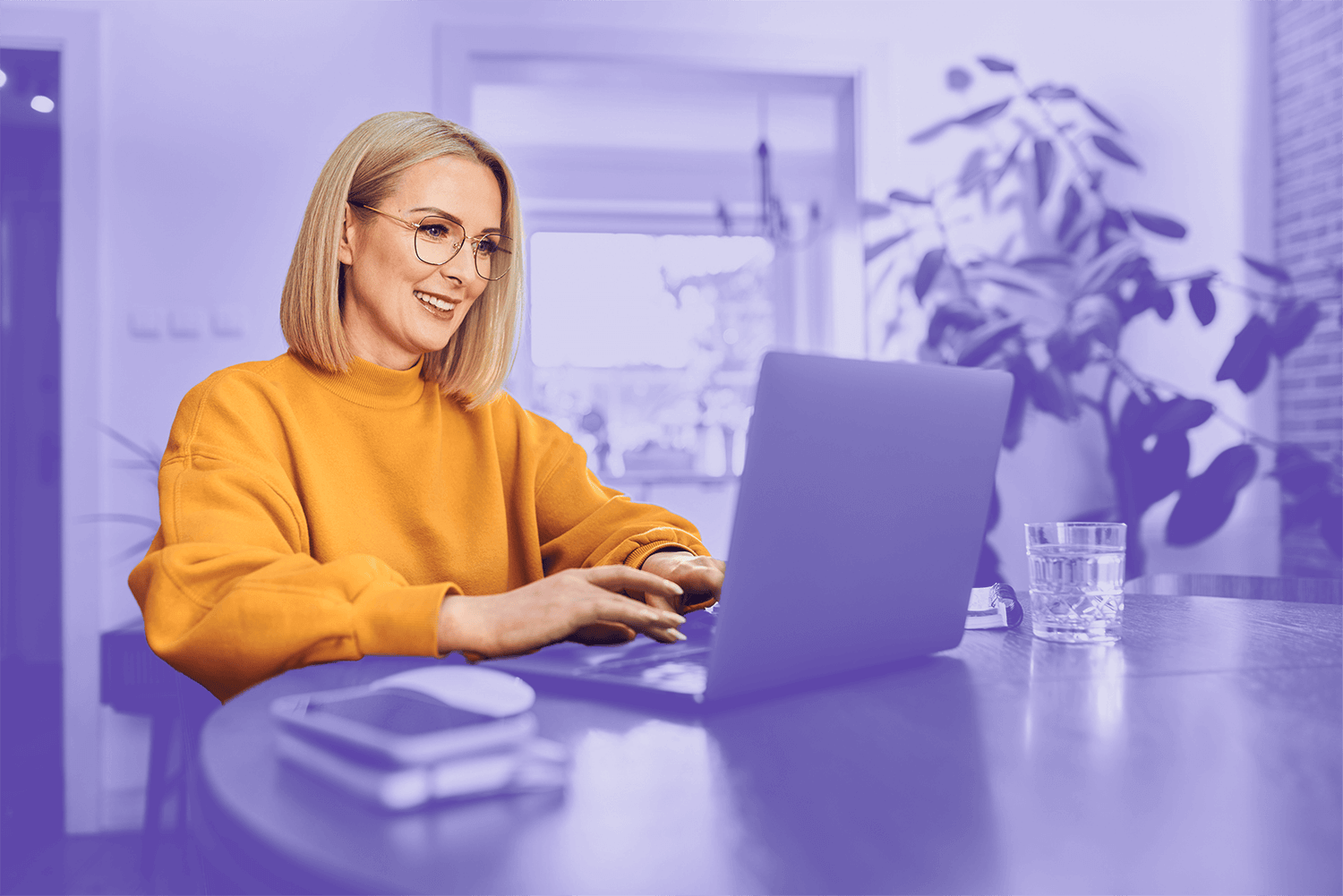 Smiling woman in a yellow sweater working on a laptop at a table in a bright home environment.