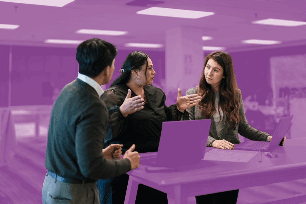 Three coworkers engaged in a discussion around a standing desk with laptops in a modern office.