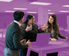 Three coworkers engaged in a discussion around a standing desk with laptops in a modern office.