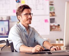 A man with brown hair and glasses sits in a wheelchair at a desk in a bright, professional workspace. He is wearing a light blue shirt and smiling while working on a desktop computer. The desk features a keyboard, mouse, and rolled-up papers, possibly design or architectural plans. The background includes blurred architectural drawings, sticky notes, and photographs pinned on a board, suggesting a creative or design-focused environment. The scene emphasizes productivity, inclusivity, and accessibility.