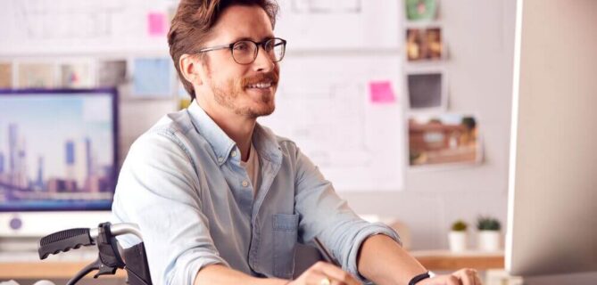 A man with brown hair and glasses sits in a wheelchair at a desk in a bright, professional workspace. He is wearing a light blue shirt and smiling while working on a desktop computer. The desk features a keyboard, mouse, and rolled-up papers, possibly design or architectural plans. The background includes blurred architectural drawings, sticky notes, and photographs pinned on a board, suggesting a creative or design-focused environment. The scene emphasizes productivity, inclusivity, and accessibility.