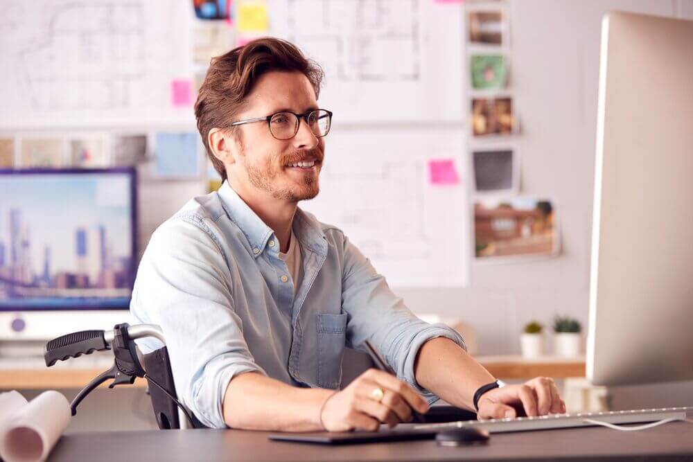A man with brown hair and glasses sits in a wheelchair at a desk in a bright, professional workspace. He is wearing a light blue shirt and smiling while working on a desktop computer. The desk features a keyboard, mouse, and rolled-up papers, possibly design or architectural plans. The background includes blurred architectural drawings, sticky notes, and photographs pinned on a board, suggesting a creative or design-focused environment. The scene emphasizes productivity, inclusivity, and accessibility.