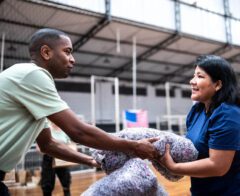 Two volunteers smiling while passing a bundle of donated blankets inside a gymnasium during a relief effort.