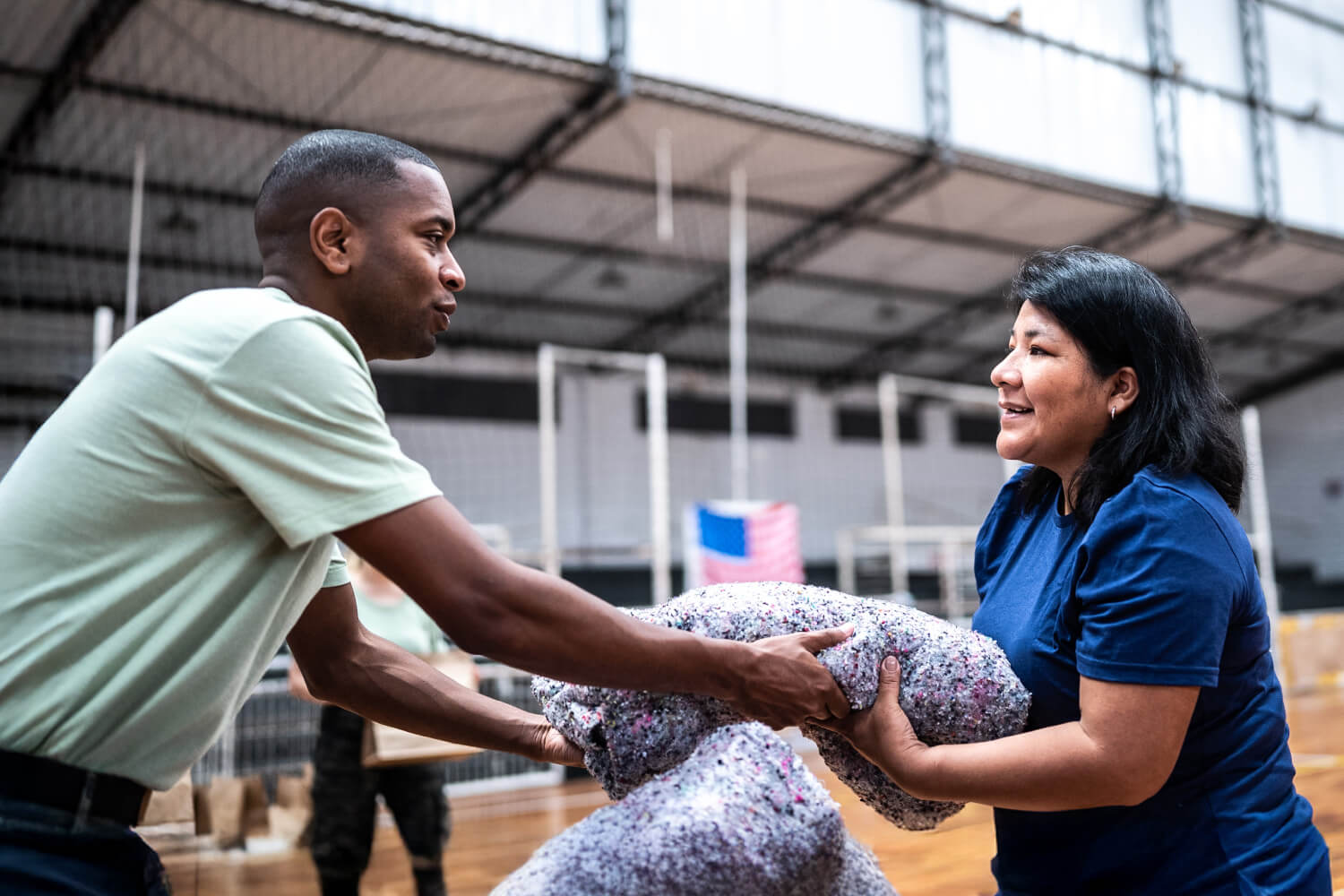 Two volunteers smiling while passing a bundle of donated blankets inside a gymnasium during a relief effort.