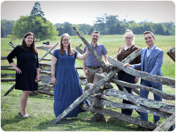Group of five adults smiling and posing by a historic wooden fence in a grassy field.