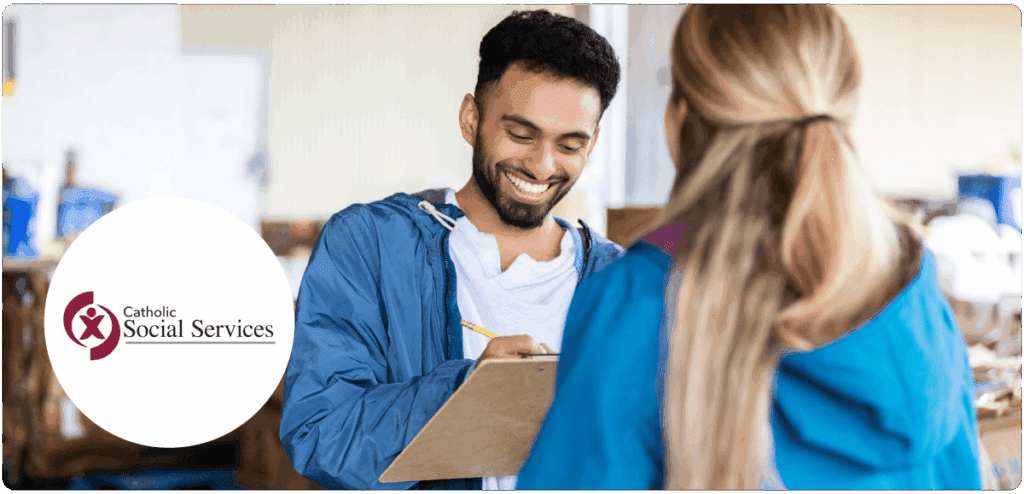 Smiling man with a clipboard talking to a woman in a blue jacket, with the Catholic Social Services logo displayed.