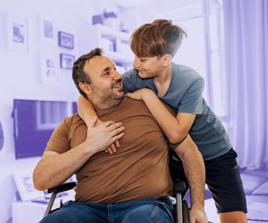 A warm interaction between a man in a wheelchair and a young boy in a living room. The man, wearing a brown t-shirt and jeans, smiles as the boy, in a blue t-shirt and black shorts, leans over to embrace him. The background features shelves, framed pictures, and a television, with a soft purple hue overlay emphasizing the subjects.