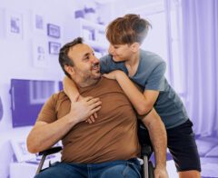 A warm interaction between a man in a wheelchair and a young boy in a living room. The man, wearing a brown t-shirt and jeans, smiles as the boy, in a blue t-shirt and black shorts, leans over to embrace him. The background features shelves, framed pictures, and a television, with a soft purple hue overlay emphasizing the subjects.