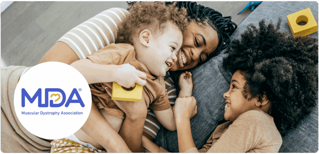 Three people lying closely together on a soft surface, smiling warmly. A child in the center holds a yellow block and is embraced by an adult on the left. Another child on the right looks at the central child. The lower left corner features the MDA® logo with the text "Muscular Dystrophy Association" in blue and a yellow heart incorporated into the "D."