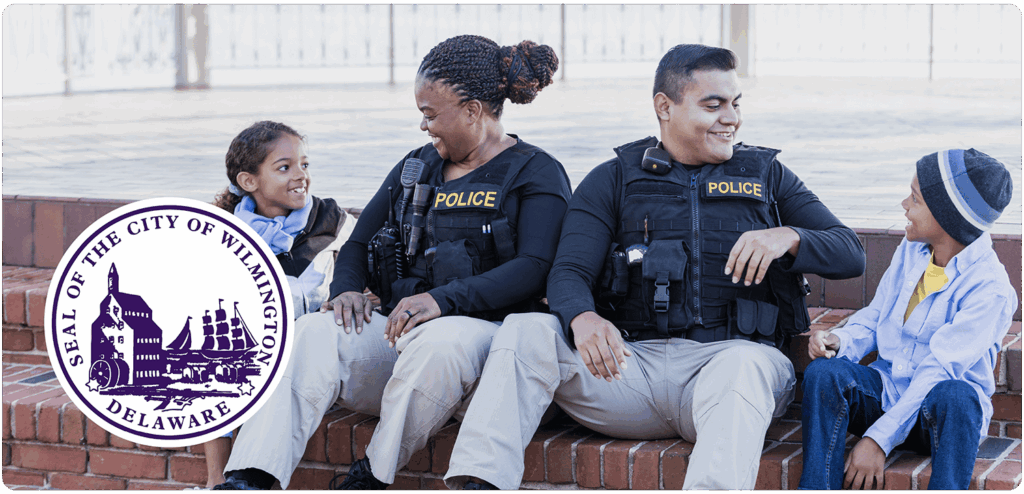 Two uniformed police officers sitting on brick steps smiling and talking with two children, with the Seal of the City of Wilmington, Delaware displayed in the foreground.