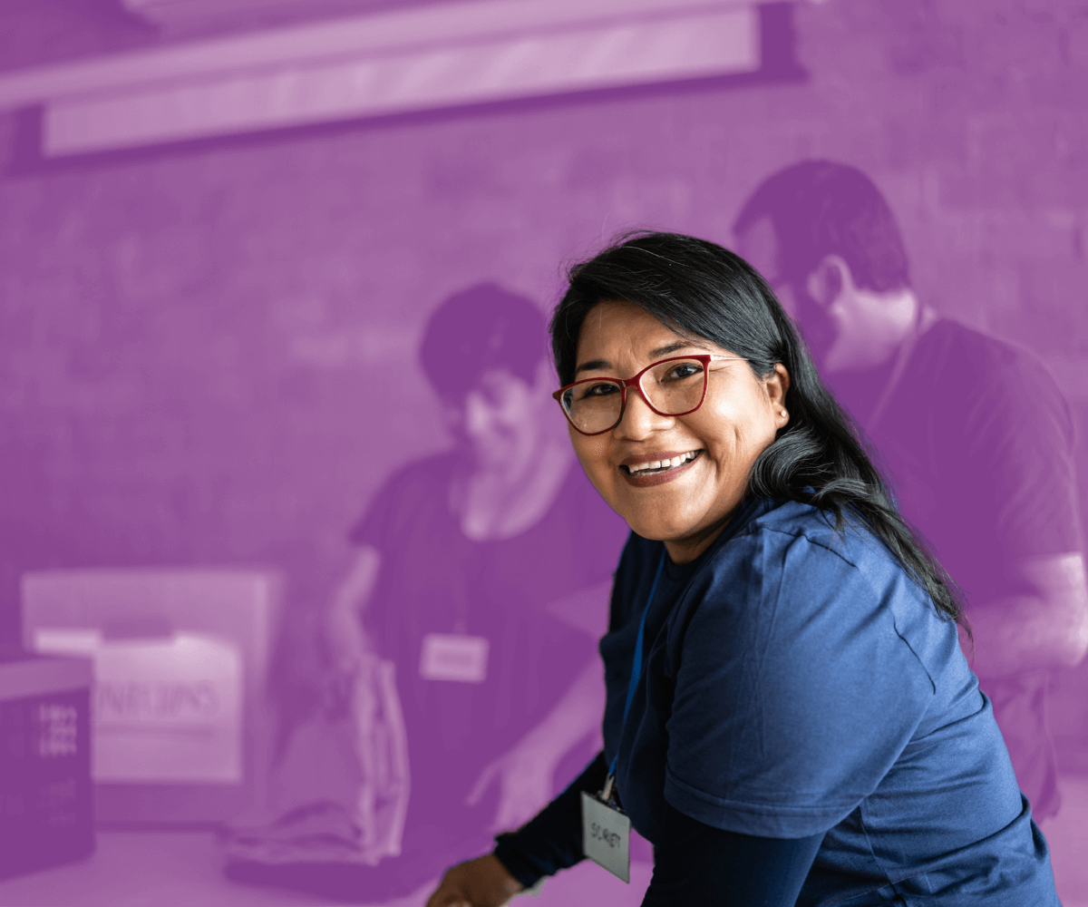 Smiling woman wearing a volunteer name tag in a community service setting with others working in the background.