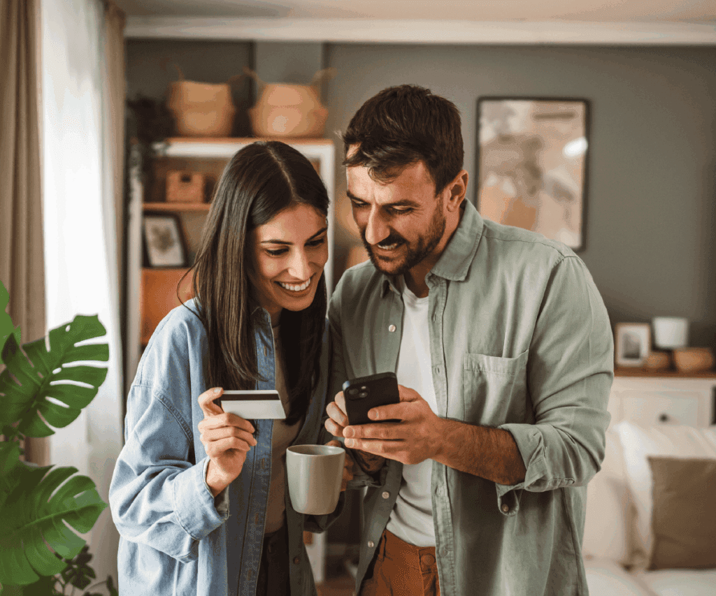 A smiling couple stands together in their living room, looking at a smartphone. The woman holds a credit card and a coffee mug, while the man points at the phone screen. They appear engaged and happy, suggesting they are making an online donation or purchase.